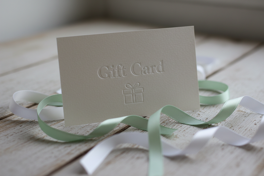 a close up picture of a gift card, That has the words Gift Card printed on it, sitting on a wooden surface with scraps of pastel green and white ribbon around the card